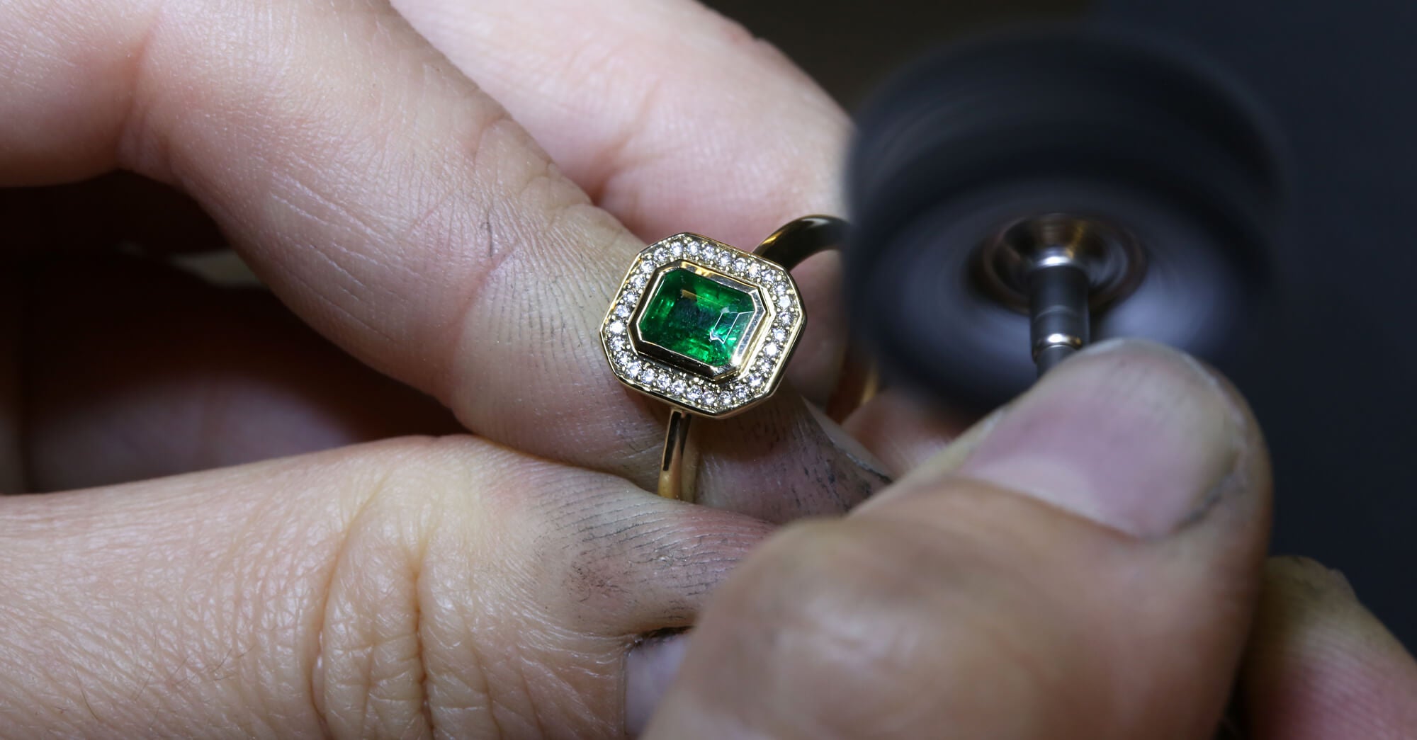 Close-up of a jeweller's hand polishing a gold ring with a green emerald gemstone with a tool.
