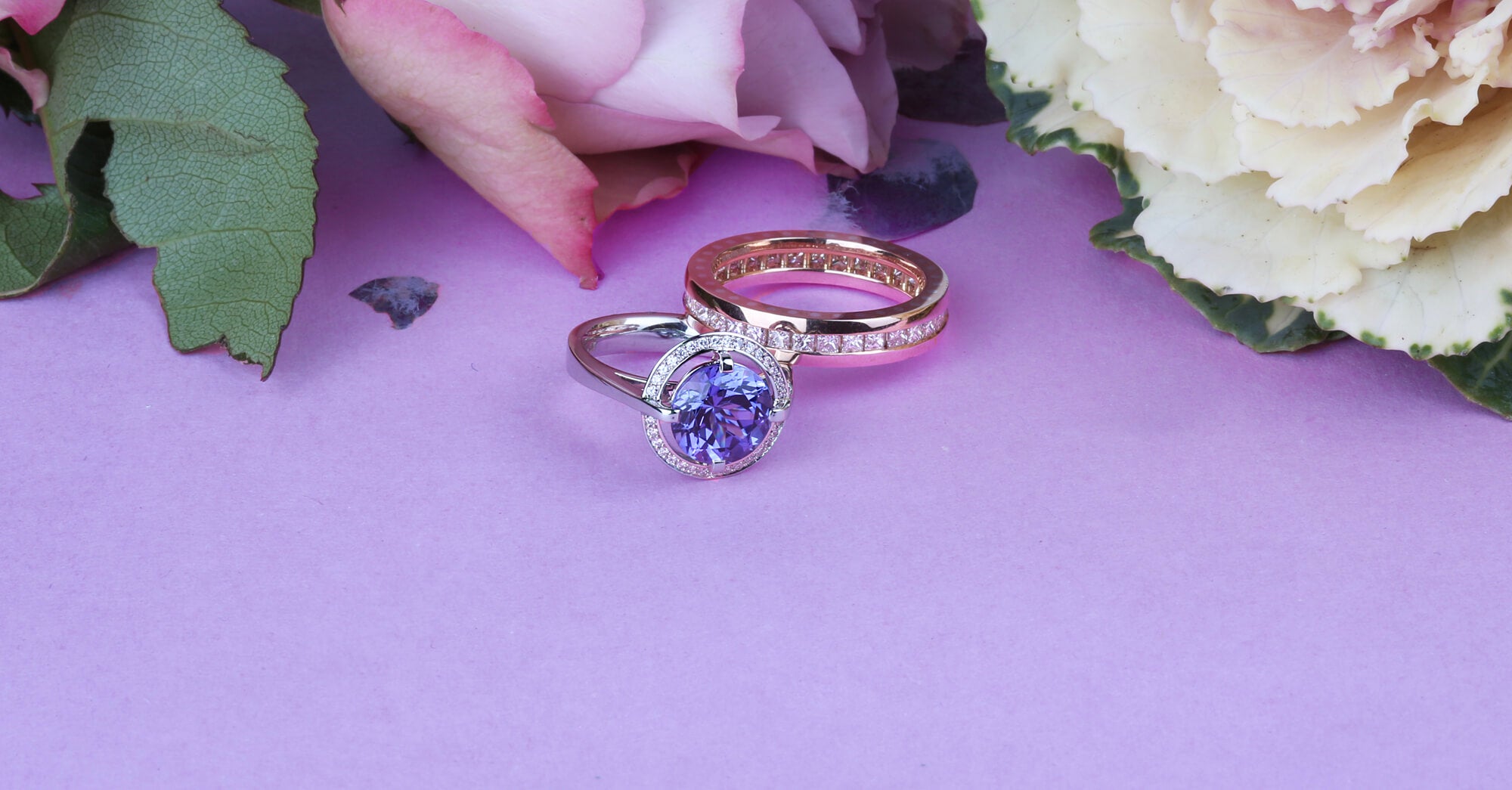 A white gold ring set with a large round brilliant cut tanzanite surrounded by pavé set diamonds sits next to a rose gold diamond set eternity ring