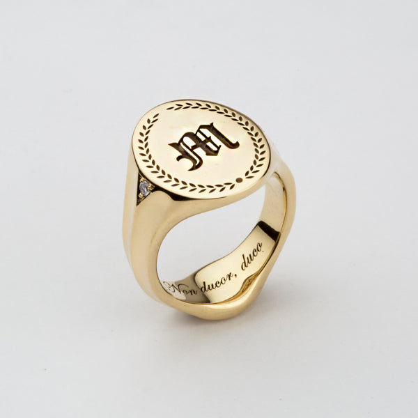 A yellow gold oval Signet ring with an initial and a pattern around it engraved at the top. Additional engraving has been placed on the inside of the band and a diamond is set on the side.