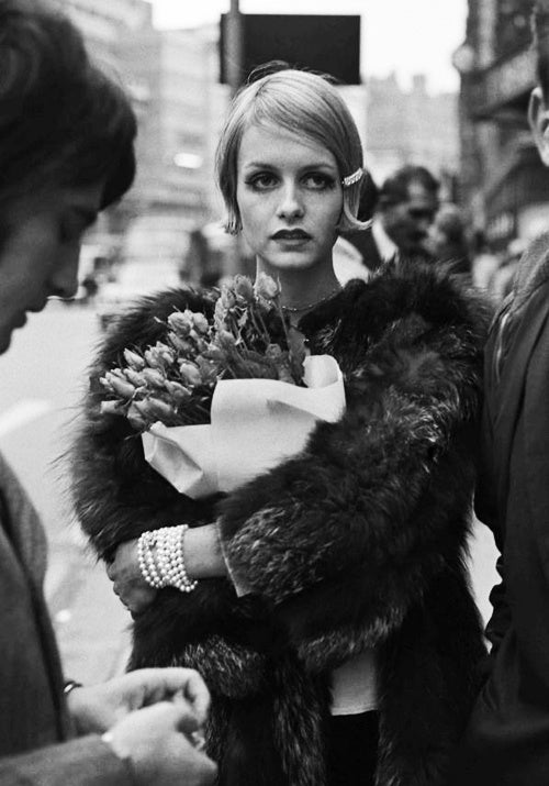 Black and white street photo of Twiggy in a fur coat holding tulips, wearing a multi-strand pearl bracelet