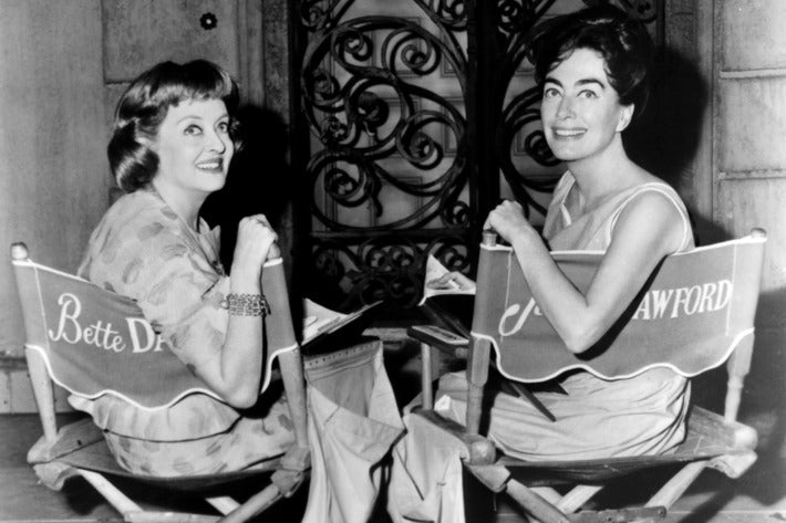 Two Hollywood actresses smiling in director's chairs bearing their names on a film set, black and white