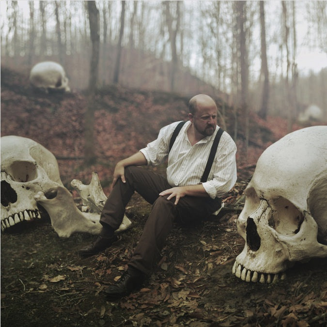 Surreal photograph of a man in braces sitting among giant human skulls on a woodland floor