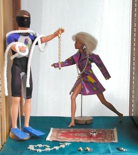 Two dolls in a shop window display, one handing a gold chain to a diver, with jewellery laid out on a small rug