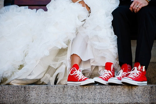 Bride in a ruffled white gown and groom in black suit sitting side by side on steps in matching red trainers