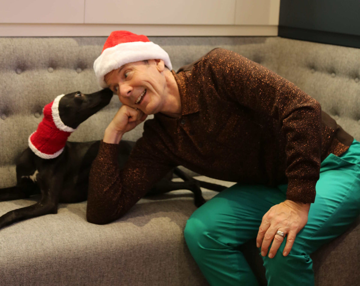 Man in a Santa hat and glittery brown shirt leaning on a sofa beside a black dog in a knitted Christmas hat