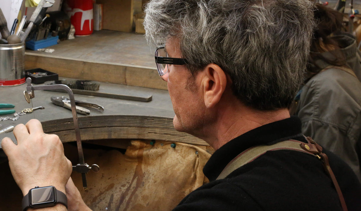 Grey-haired jeweller in glasses working at the bench peg in our workshop, tools laid out beside him
