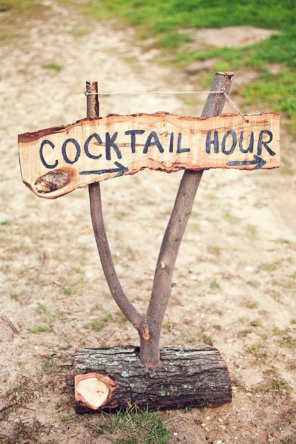 Rustic wooden sign reading 'Cocktail Hour' with arrows, nailed to a forked branch outdoors