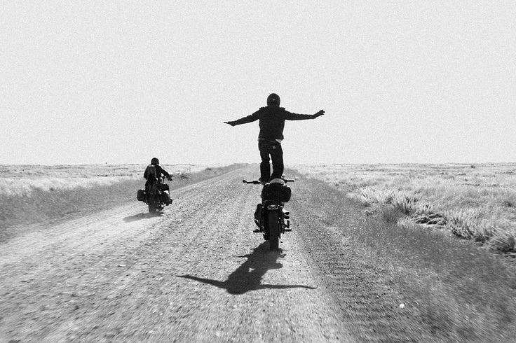 Black and white photo of a rider standing on his moving motorbike, arms outstretched, on a dirt road