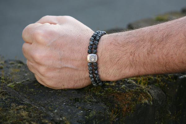 Man's clenched fist wearing a double strand bracelet of snowflake obsidian beads with a silver bead, on a mossy rock