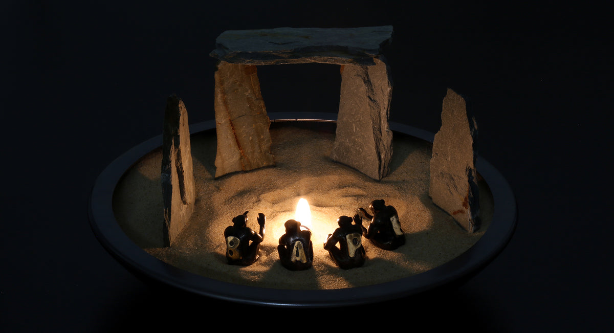 Small figures seated around a candle inside a miniature stone circle on sand, lit in the dark