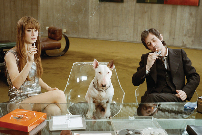 Film still of a woman in a sequin dress and a man in a suit, with a bull terrier on a clear chair between them
