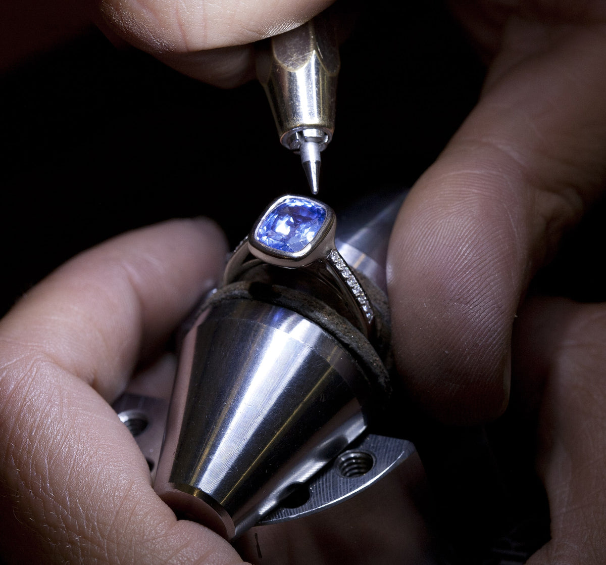 Jeweller setting a cushion-cut blue sapphire in a white gold ring with diamond shoulders on a ring clamp