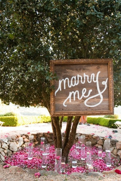 Wooden sign reading "marry me" hung on a tree above rose petals and glass candle holders