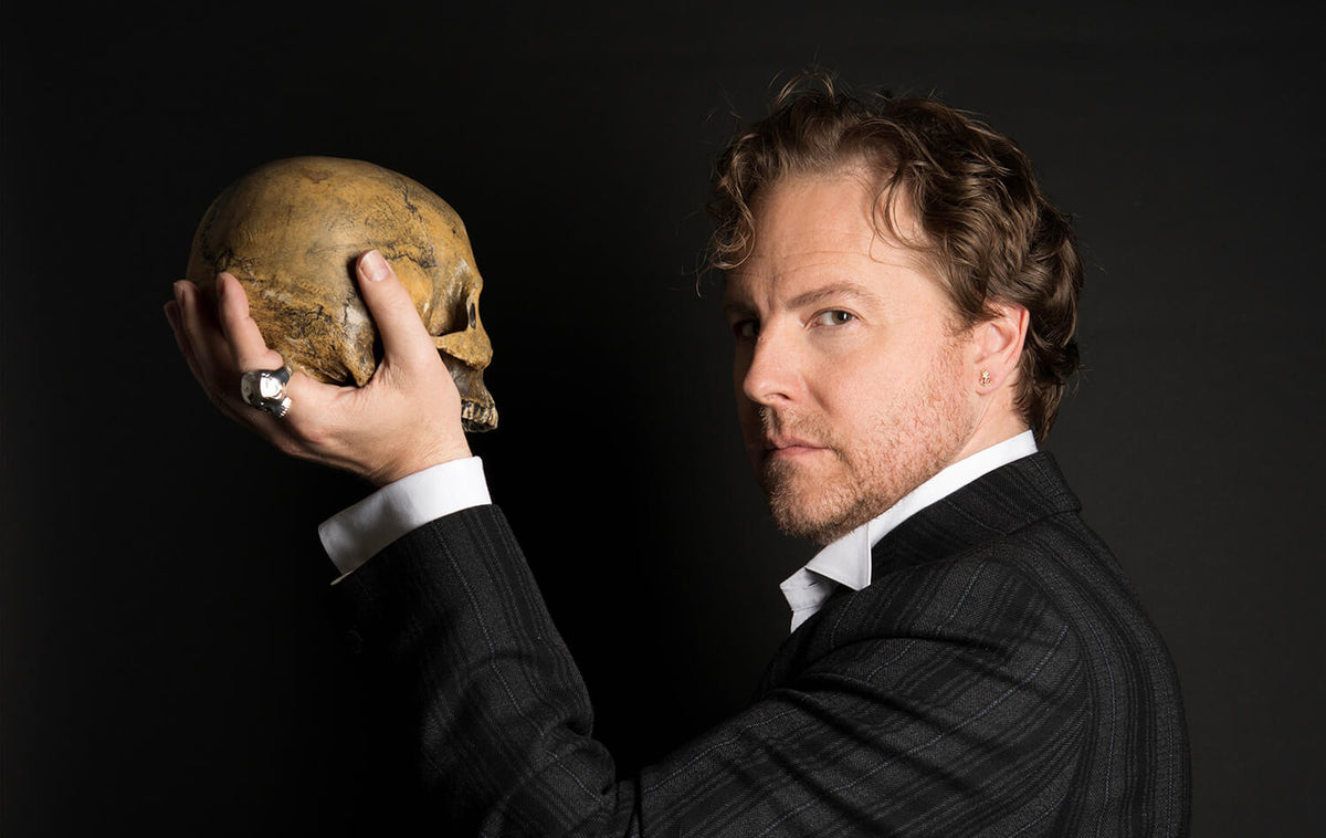 Samuel West holding a skull in his hand, wearing a large silver ring, against a black background