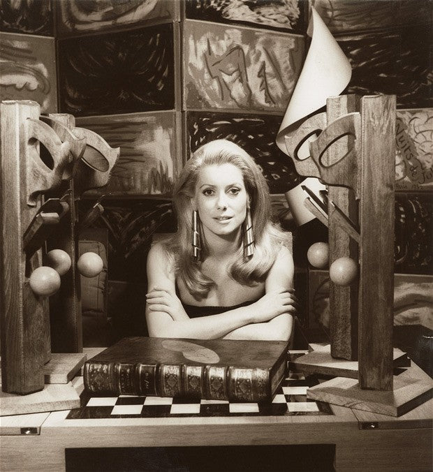 Sepia portrait of a blonde woman in long bar drop earrings, arms folded on a book among wooden sculptures