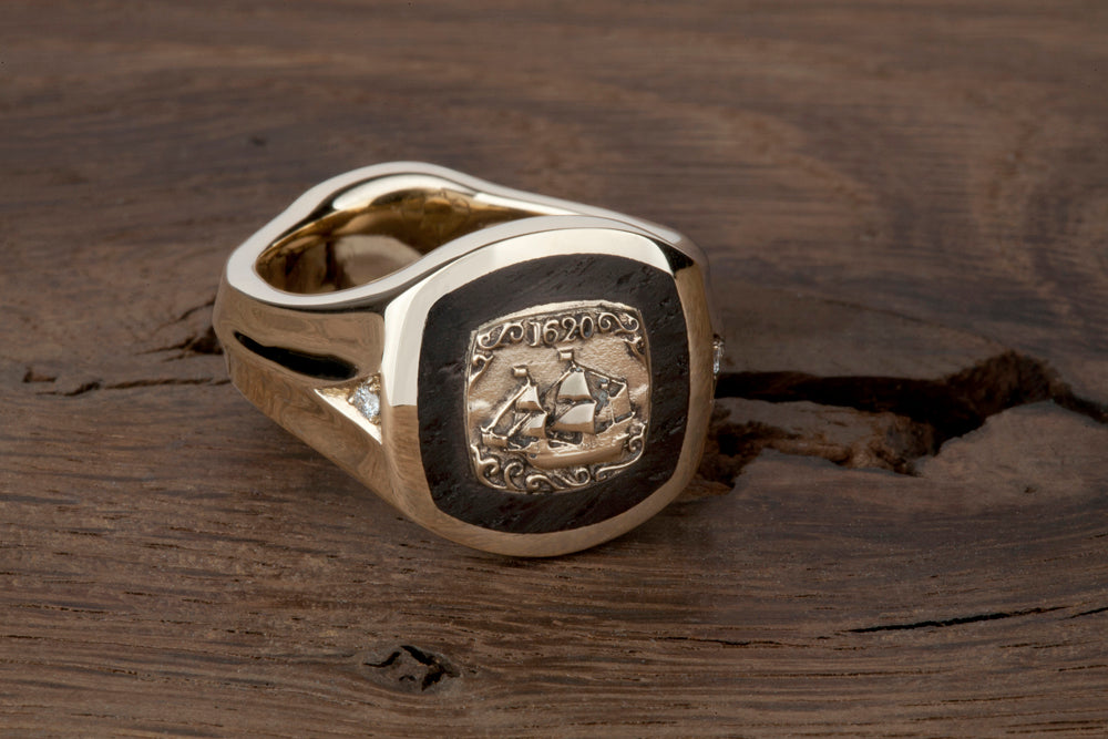 Gold Mayflower signet ring with a raised ship and 1620 on dark Thames wood inlay, resting on weathered timber