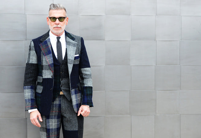 Grey-haired man in sunglasses wearing a patchwork tartan wool suit and tie against a grey tiled wall