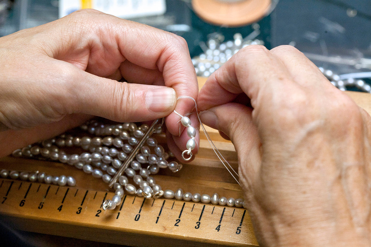 Hands knotting a strand of freshwater pearls on silk over a measured wooden pearl-stringing board