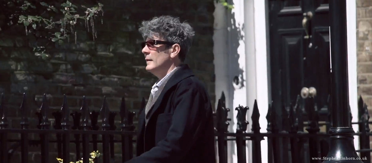 Grey-haired man in sunglasses and a dark coat walking past black railings on a sunny London street