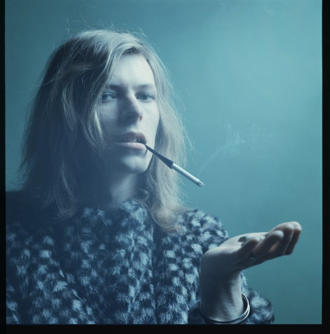 David Bowie with long hair and a cigarette in a checked coat, a silver bangle on his wrist, against teal