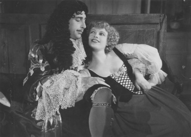 Black and white film still of a couple in 17th-century costume, a man in a long curled wig and lace