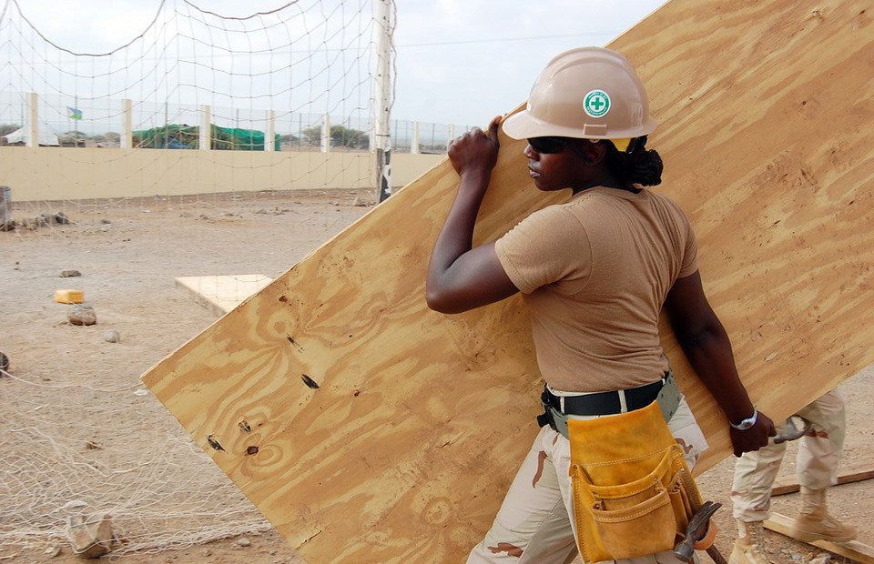 Woman in a hard hat and tool belt carrying a large sheet of plywood across a building site