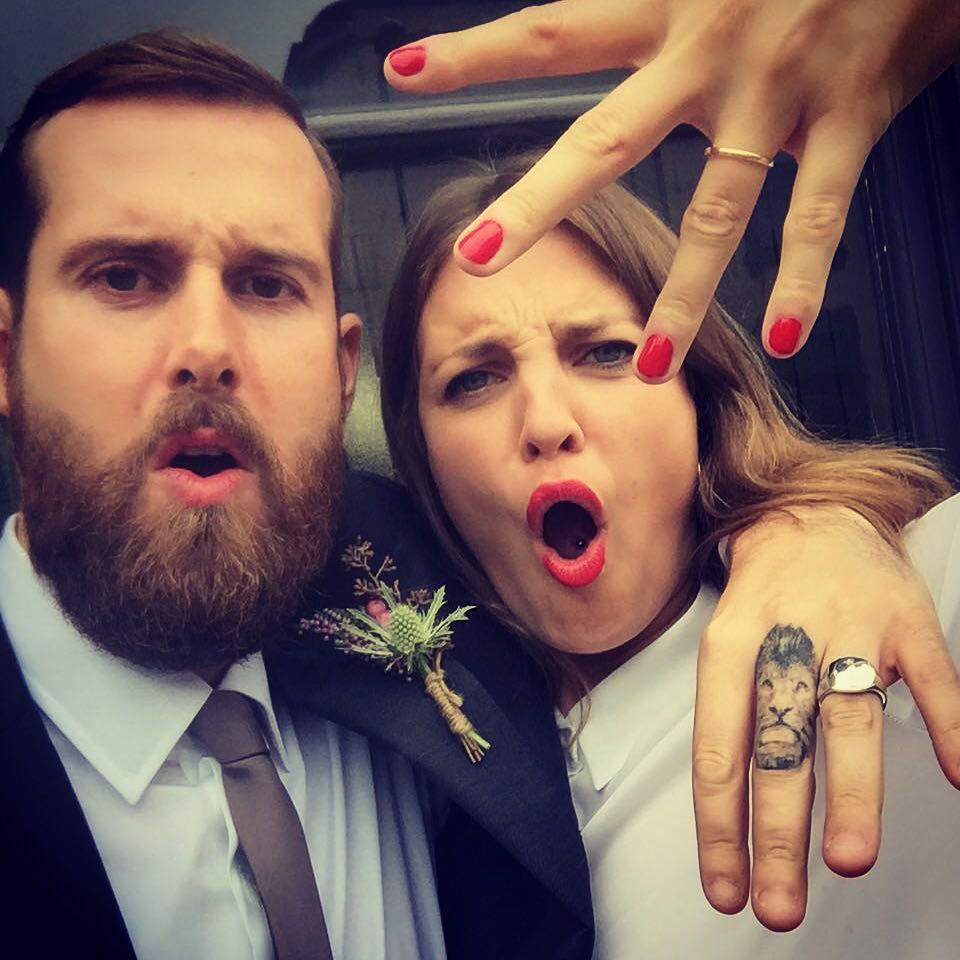 Couple on their wedding day pulling faces, the bride showing her gold wedding band and a silver ring
