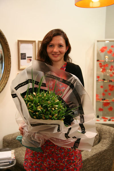 Smiling woman in a red floral dress holding a large wrapped bouquet of white tulips in our shop