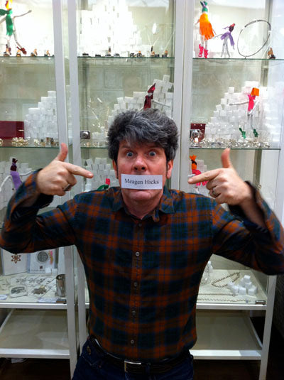Man with a name label reading 'Meagen Hicks' taped over his mouth, pointing at it in front of jewellery cabinets