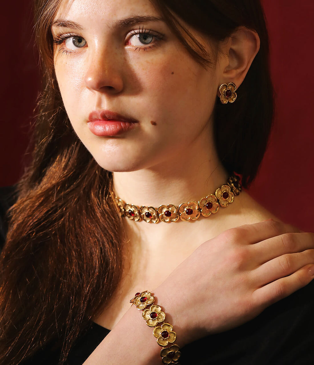 Girl wearing a gold and red gem bracelet, necklace and earrings. The jewellery design has gold interconnected flowers with red ruby's in their centres.