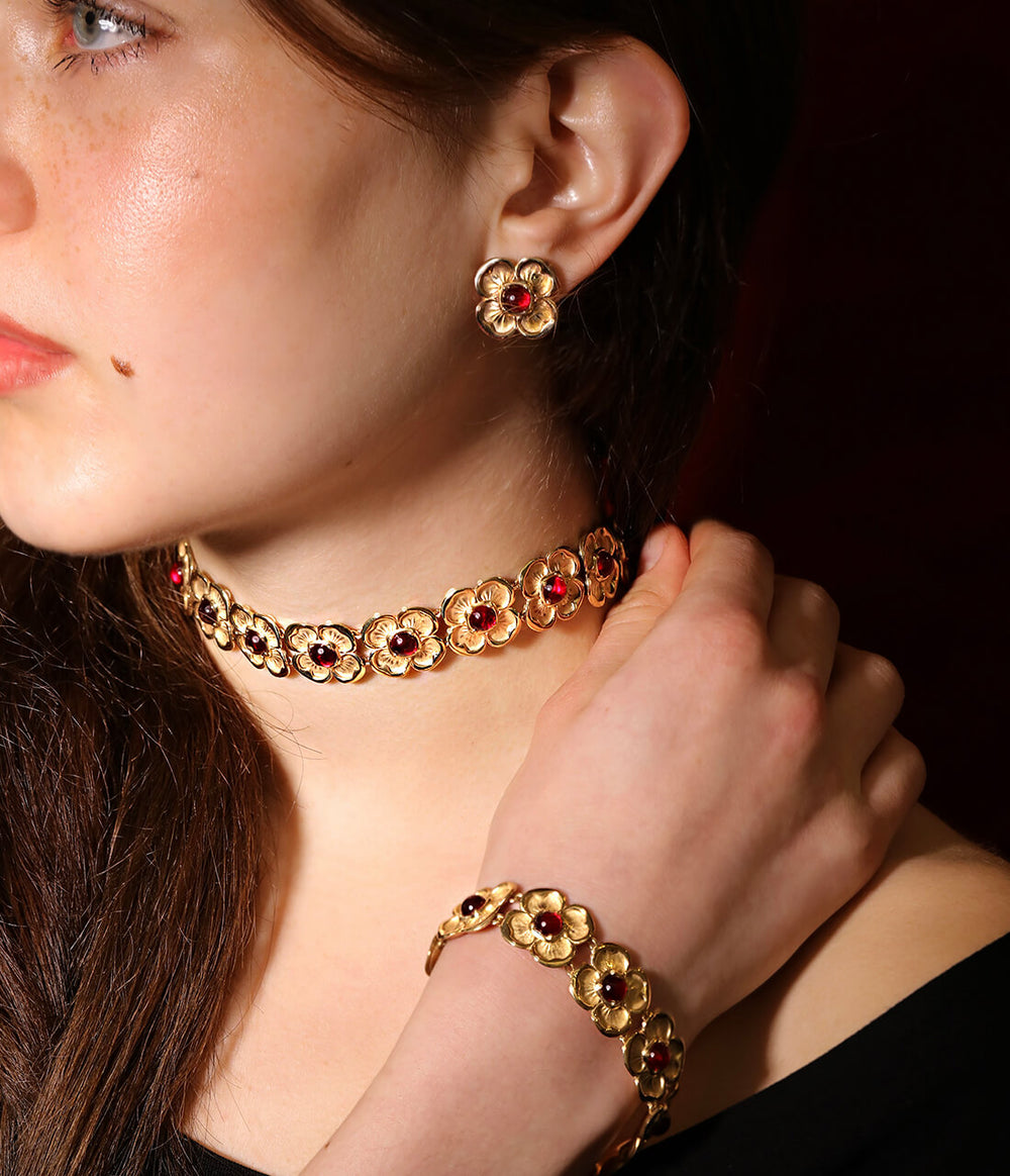 A girl wearing yellow-gold flower earrings with four-petals, each one framing a red ruby. She also wears a matching choker and bracelet made of the same interlocking gold flowers