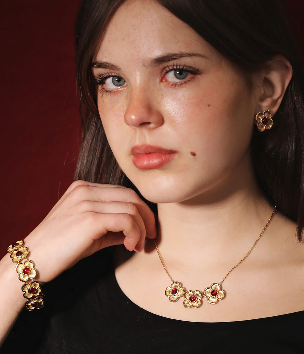 A girl wearing a yellow-gold necklace on a delicate chain with three four-petal flowers, each one holding a red ruby. She also wears a matching bracelet.