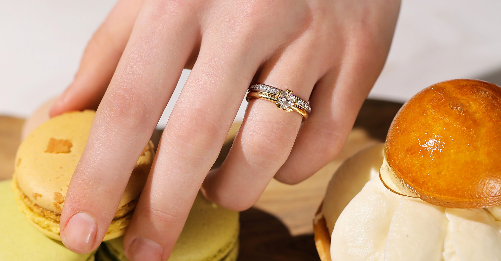 A woman's hand, who wears the 'Lotus' yellow gold diamond engagement ring and the 'Halo' platinum diamond wedding band is grabbing a macaron from a table full of desserts.