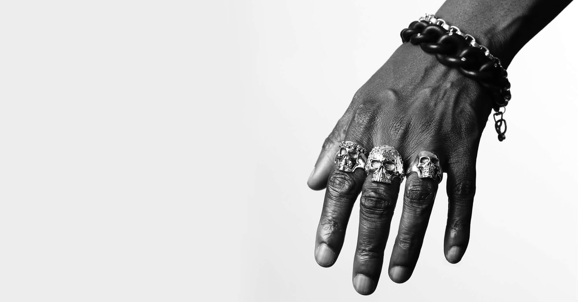 A man's hand wearing a black and silver bracelet and three skull rings on his fingers.