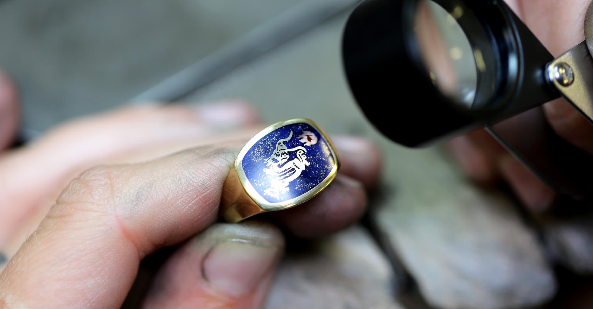 A yellow gold signet ring with blue enamel and a dragon design is being looked at by a jeweller in the workshop through a magnifying glass