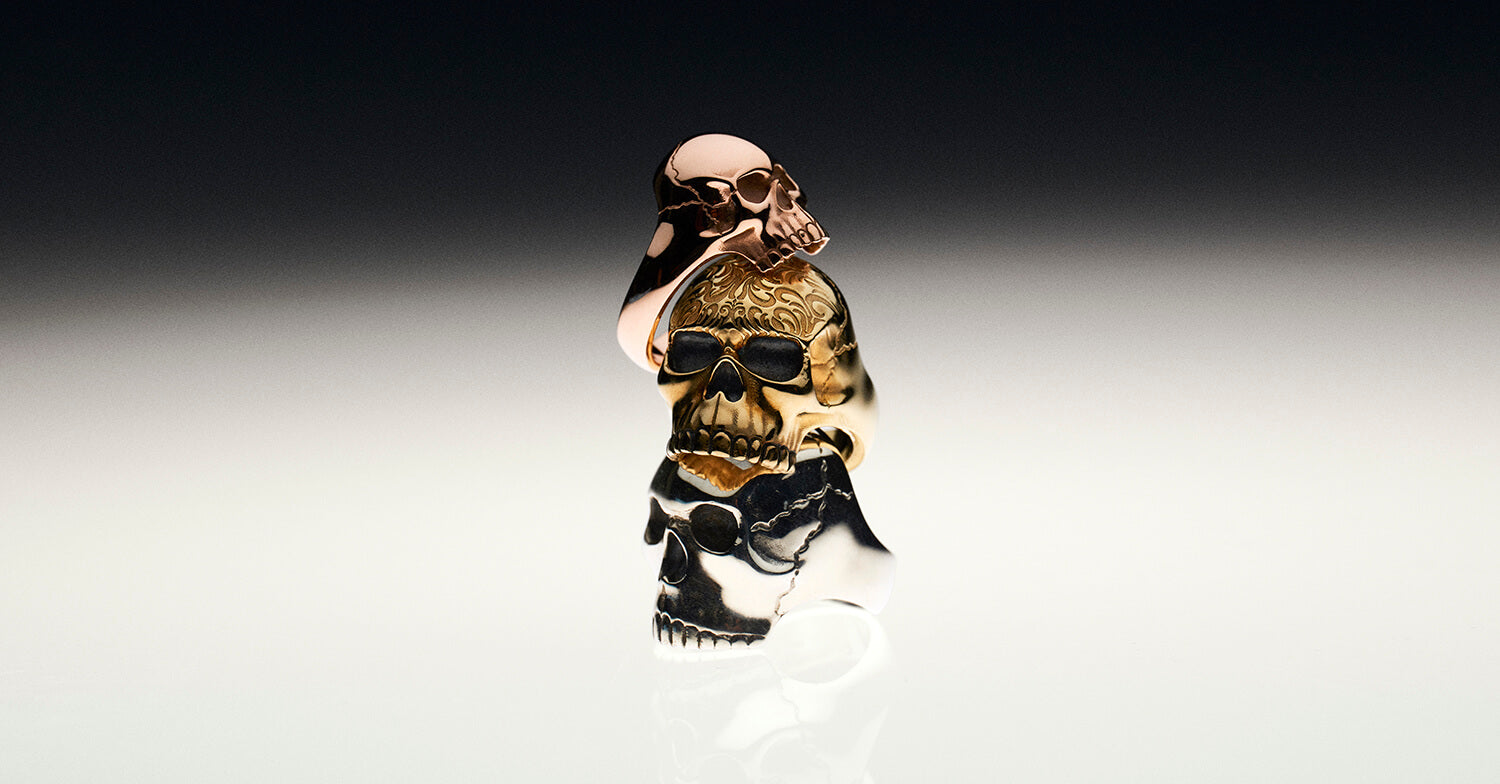 A rose gold skull ring on top of a yellow gold skull ring with a tattoo design on its head, on top of a silver large skull ring photographed on a black and white reflective background.