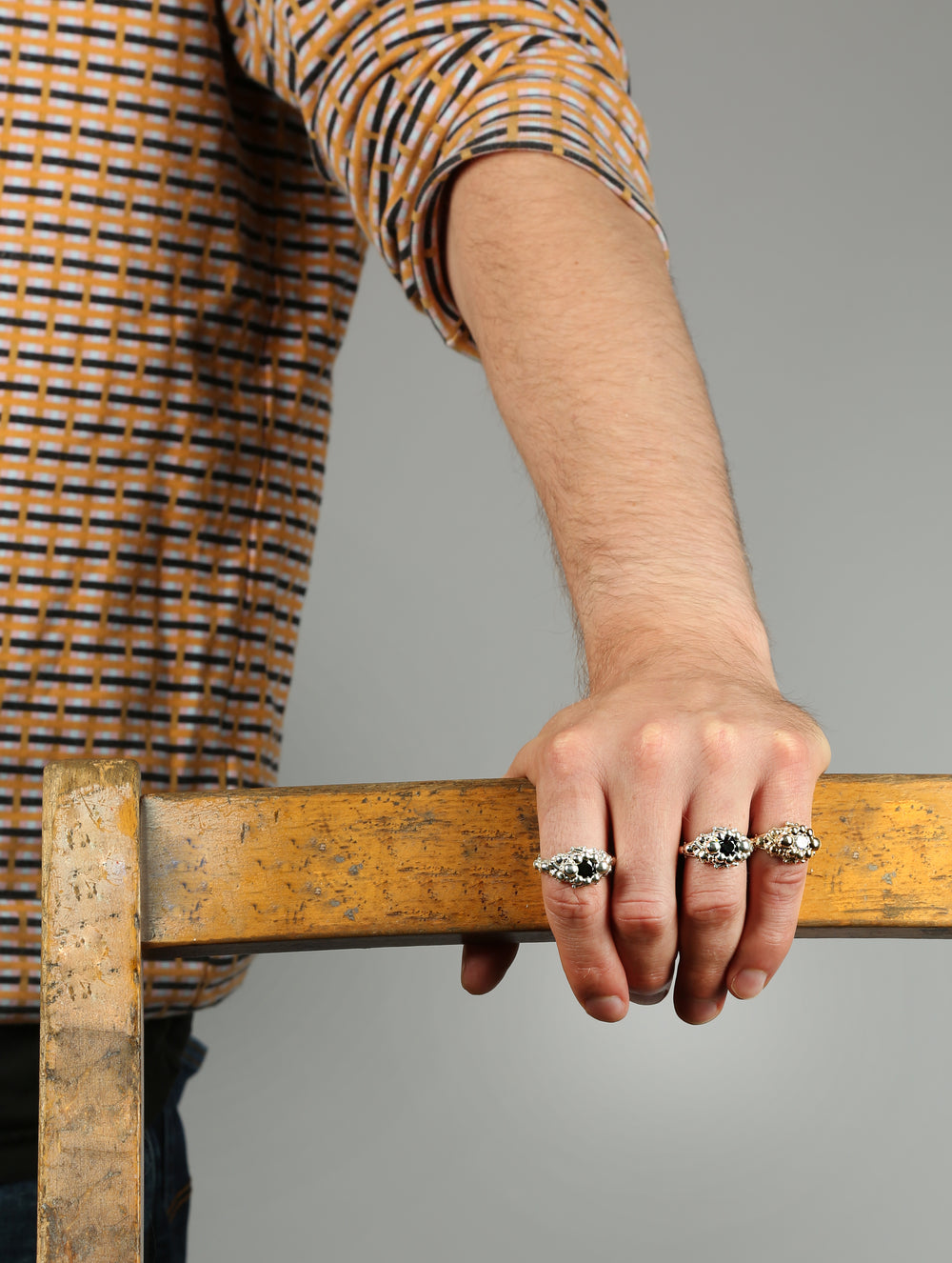 A mans hand resting on the back of  a wooden chair. He is wearing three catacomb rings with different stones set in them. He has on a patterend top.