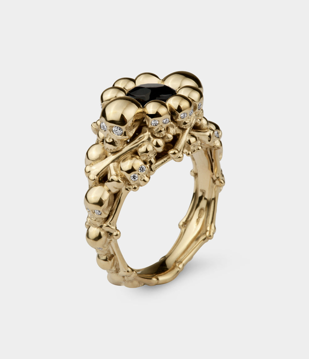 A 3/4 view of a ring made up of multiple skull and bones with a central round black diamond . The ring is in yellow gold. The small skulls have diamond eyes.