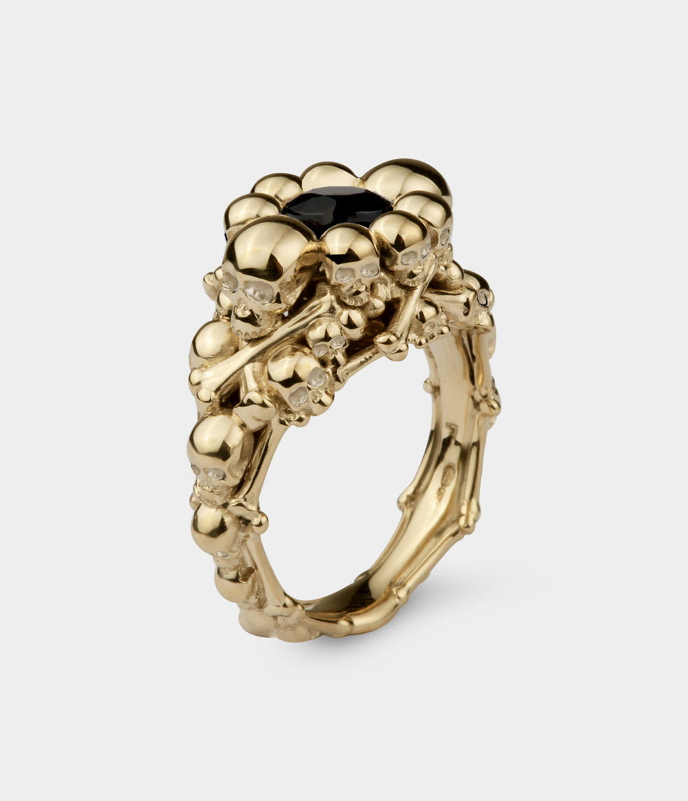 A 3/4 view of a ring made up of multiple skull and bones with a central round black diamond . The ring is in yellow gold. 