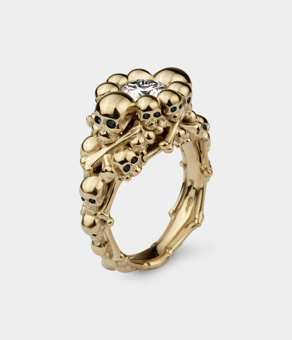 A 3/4 view of a catacomb ring made up of multiple skull and bones with a central round moissanite. The ring is in yellow gold. The small skulls have been set with black diamond eyes.