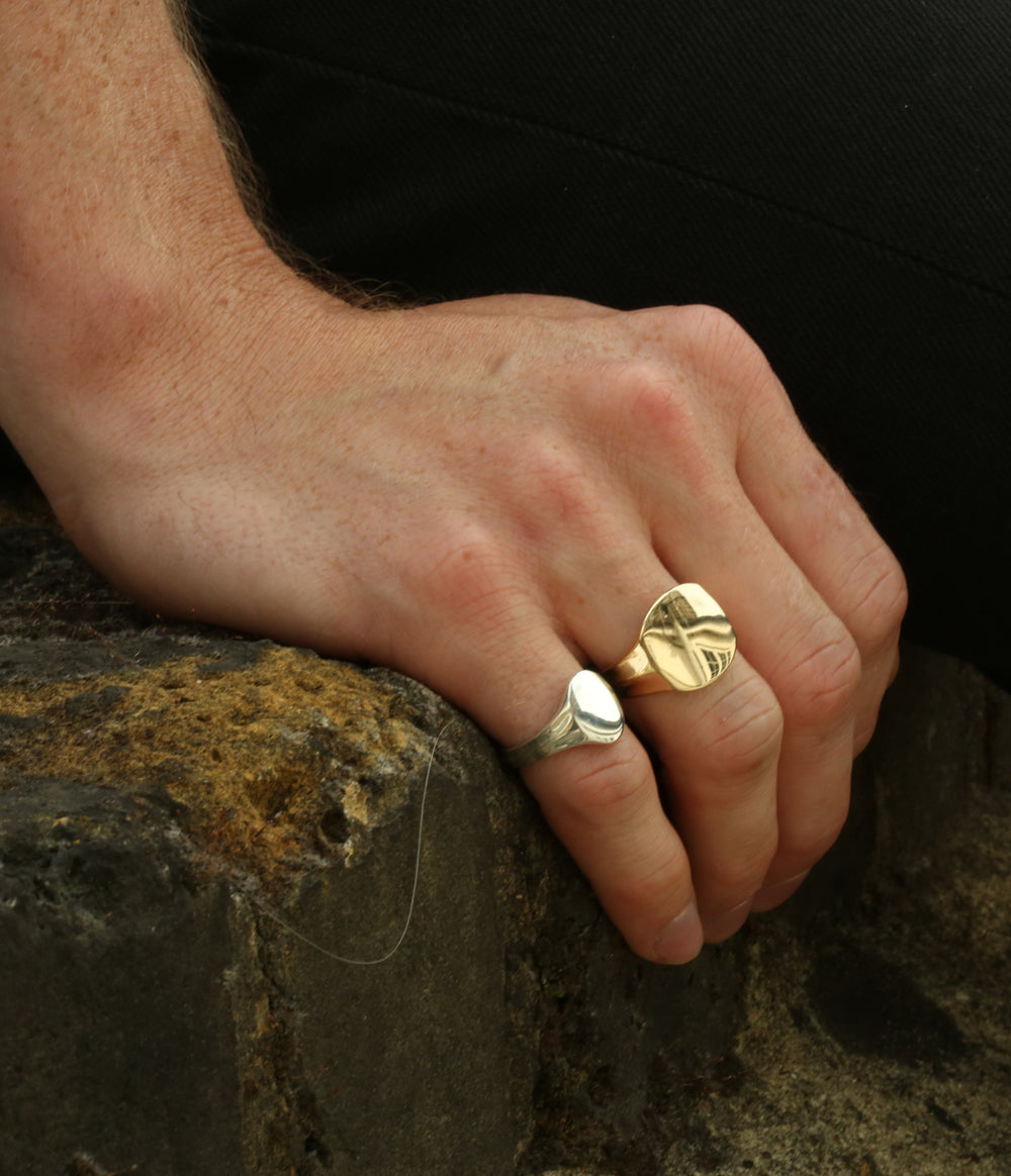 a mans hand on a rock showing the small oval signet ring in yellow gold and a yellow gold signet ring.